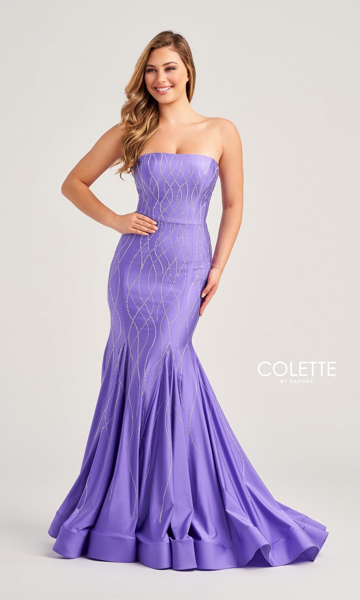 A woman smiles against a plain background in the Colette Strapless Long Glitter Prom Dress CL5106—a fitted lavender mermaid gown with silver glitter print that flares out at the bottom.
